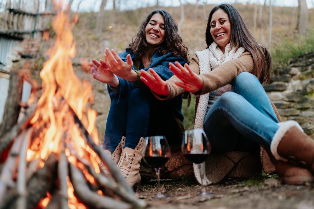 Young smiling women warming next to the fire. Glasses of red wine. Campfire, outdoors activities, relaxation, togetherness concept.